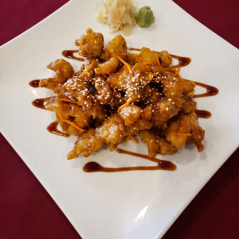 Orange chicken with sesame seeds, carrot strips, and a drizzle of sauce on a white plate.