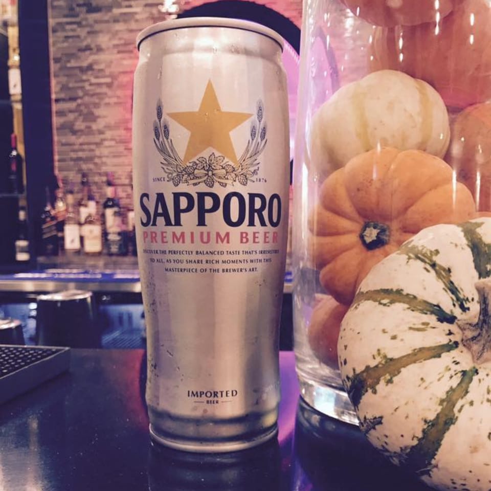 A Sapporo beer can on a bar next to a glass vase filled with small pumpkins and gourds.