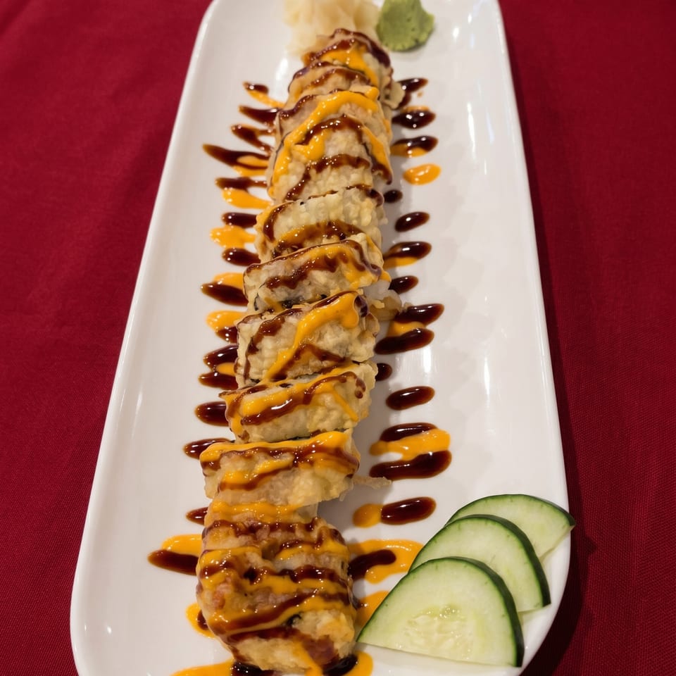 Tempura sushi roll drizzled with orange and brown sauces, served with cucumber slices on a white plate.
